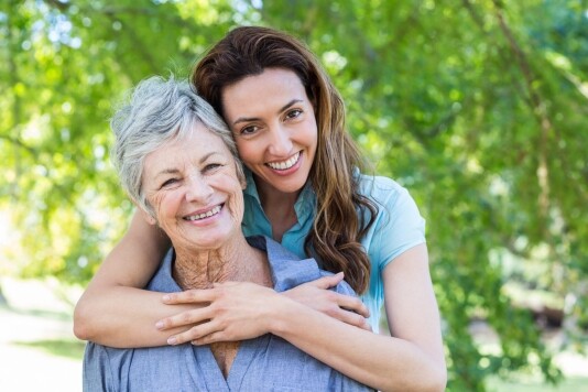 An older woman and a younger woman smile outdoors on a beautiful day in May 2024. The younger woman has her arms wrapped around the older woman's shoulders from behind as they stand in front of lush local green foliage.