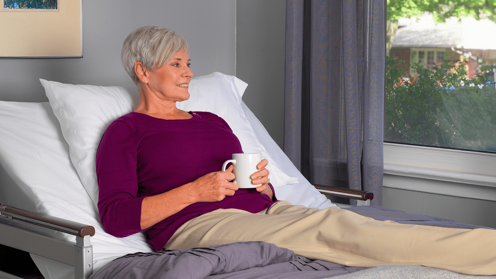 An older woman with short gray hair smiling, sitting up in bed with a cup, looking out a window in Apr2024.