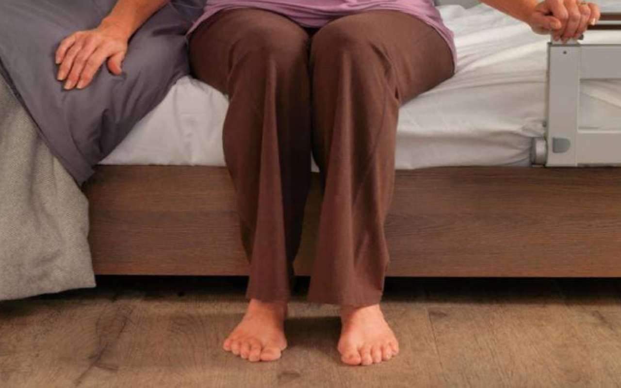 A person sits on the edge of a bed with their feet touching the floor. They are wearing brown pants and a purple top, with one hand resting on the bed and the other on a bed rail.