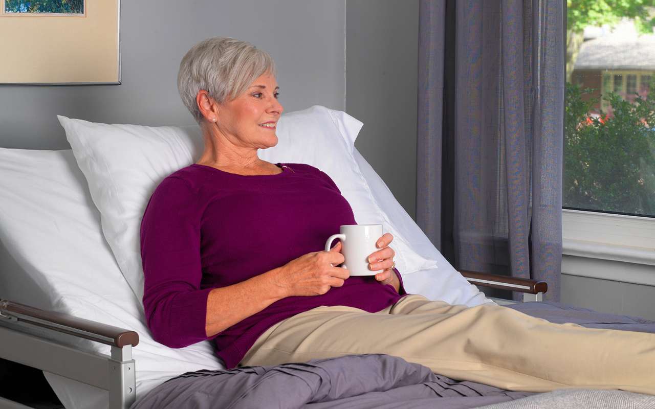 An elderly woman with short gray hair is sitting up in a bed, holding a white cup, and looking out the window. She is wearing a purple sweater and beige pants.