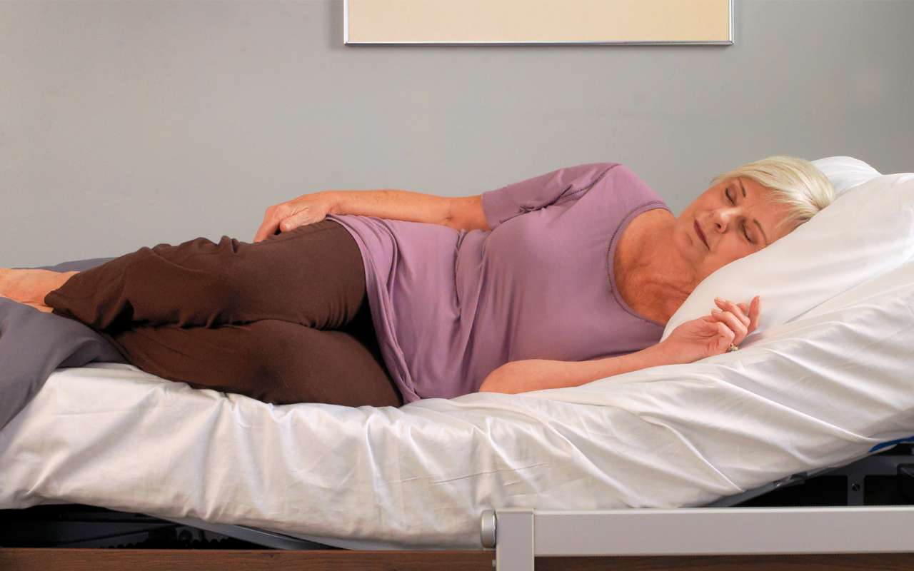 An older woman in a purple top and brown pants sleeps on her side in a bed with white sheets and a gray blanket.