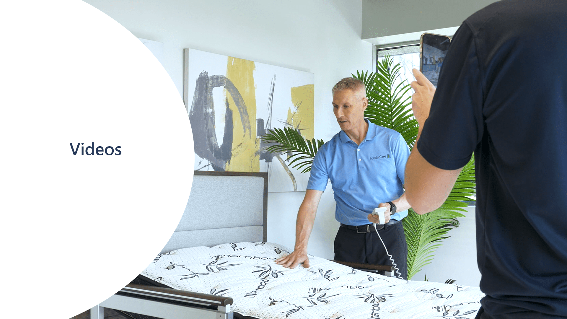 A man demonstrating a product on a bed while another person films him, capturing every detail for their videos. The word "Videos" is displayed in a white circular overlay on the left side of the image.