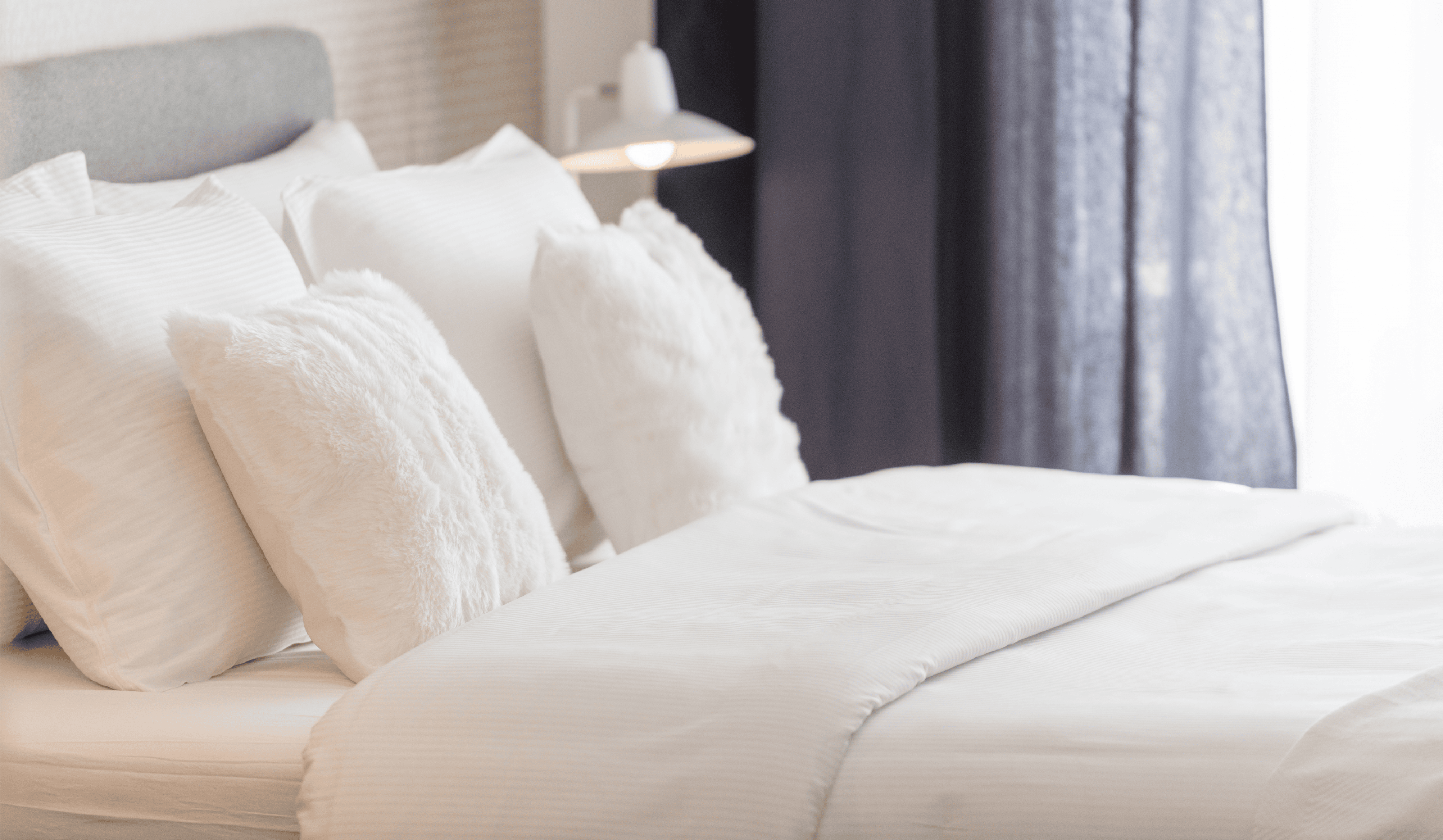 A neatly made bed with fresh, new bedding featuring white pillows and sheets, next to a small bedside lamp and dark curtains.