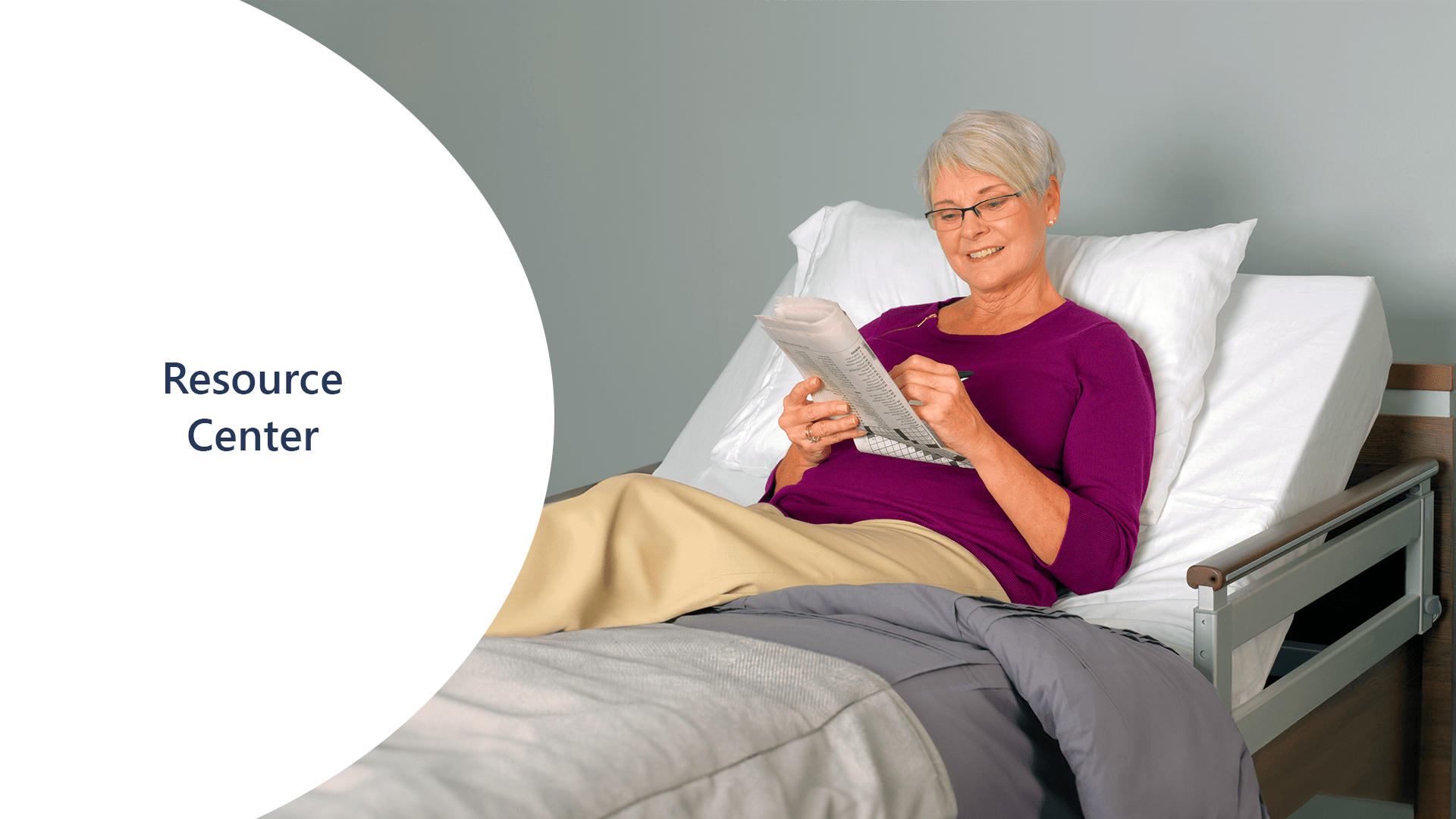 An elderly person in a purple top and beige pants is reading about current events in a newspaper while reclining on a bed in a light-filled room.