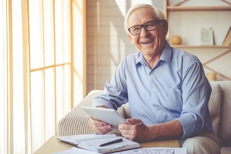 An older man with glasses smiles as he sits on the couch, holding a tablet. Papers and a pen lie about on the table in front of him.