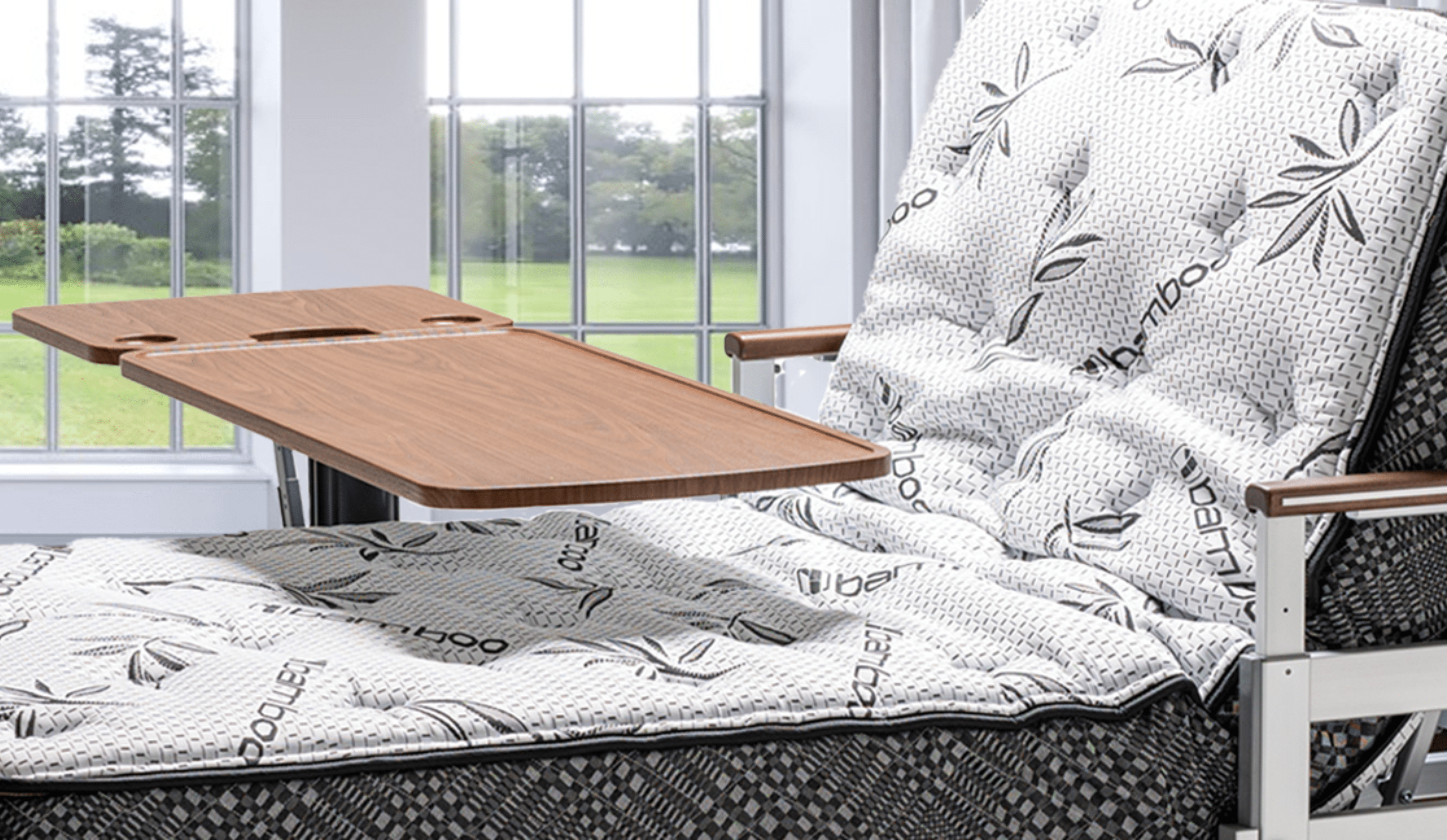 An adjustable hospital bed with a wooden tray and new accessories stands in a sunlit room, featuring patterned bedding.