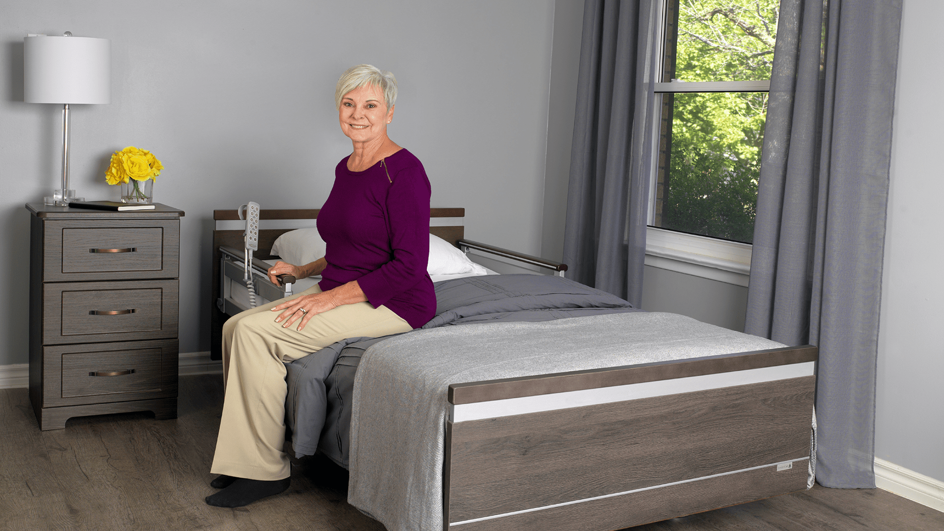 A mature woman with short gray hair, wearing a purple top and beige pants, sitting on the edge of a bed in a neatly decorated room with a window.