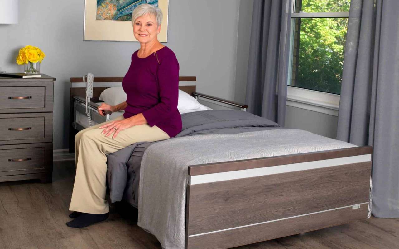 An older woman sits on the edge of a bed in a well-lit bedroom with grey and wood tones. She holds onto a bed assist rail, which is attached to the side of the bed.