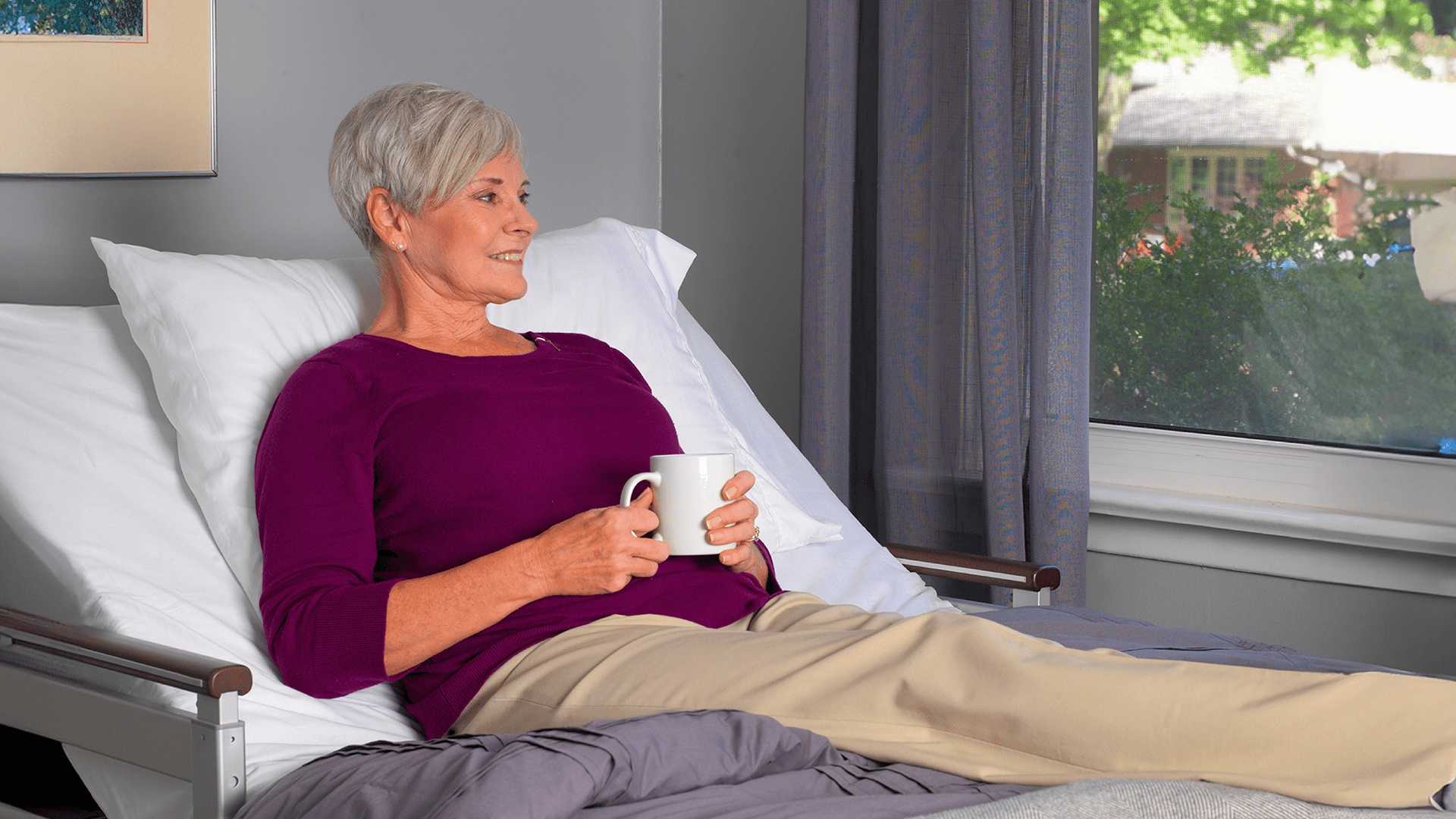 Senior woman sitting in a hospital bed, holding a white mug, smiling and looking out the window.