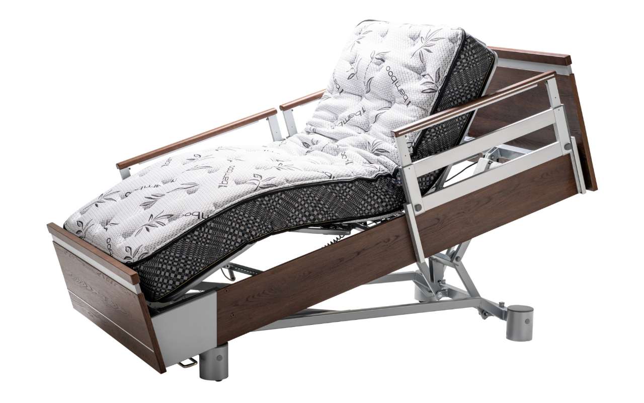 A hospital bed with a wooden frame, equipped with side rails and an adjustable head section, is angled upwards. The bed is made with patterned bedding.