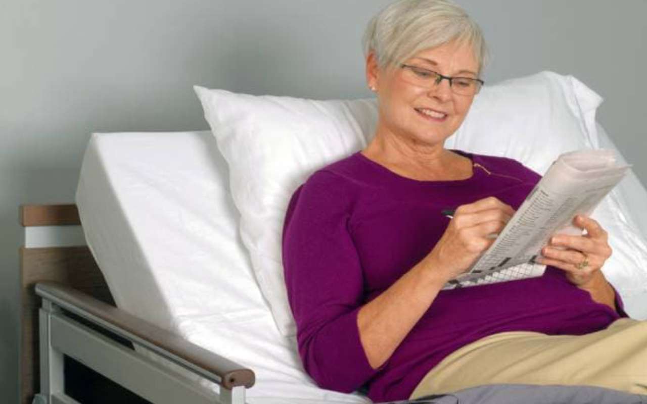 An elderly woman wearing glasses and a purple sweater is sitting up in a hospital bed, smiling while writing on a newspaper.