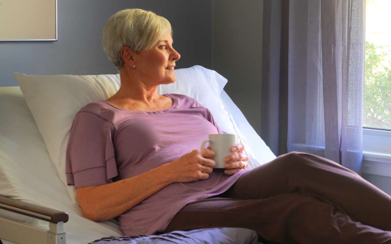 A woman with short gray hair sits in a hospital bed, holding a cup, and gazes outside through a window. She wears a purple shirt and dark pants.