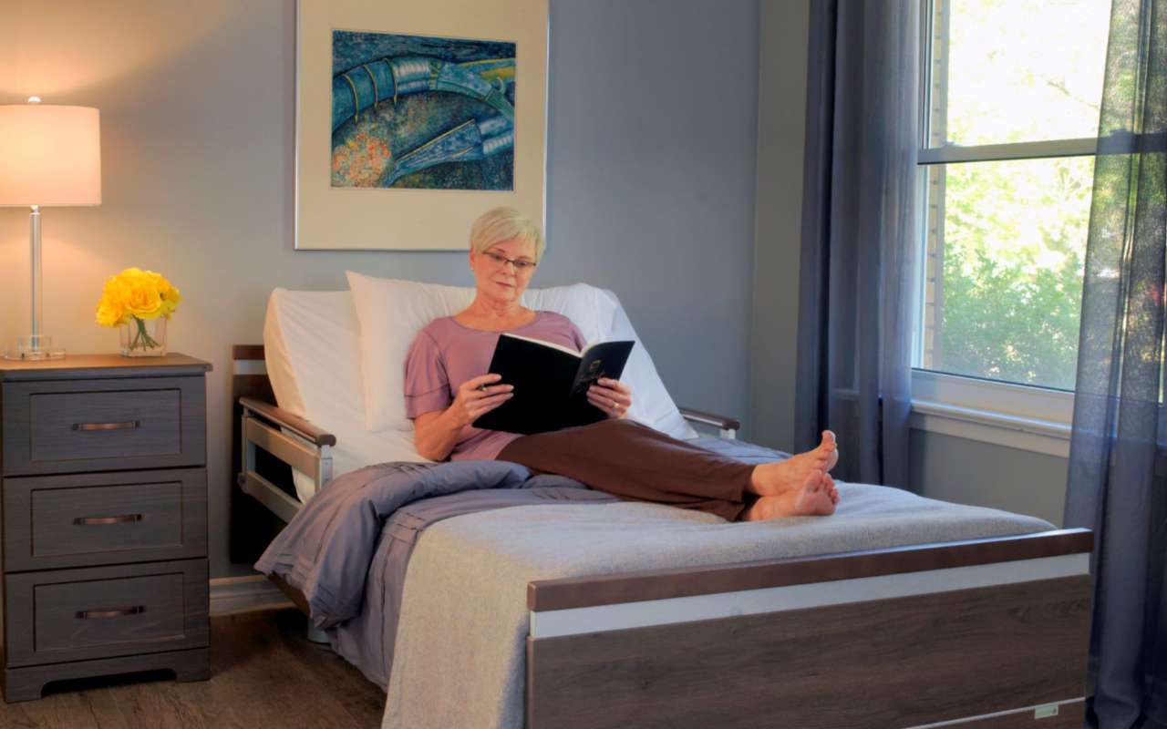 A woman with short, white hair, wearing glasses, sits in a hospital bed reading a book. The room has gray walls, a nightstand with a lamp and flowers, and a window with sheer curtains.